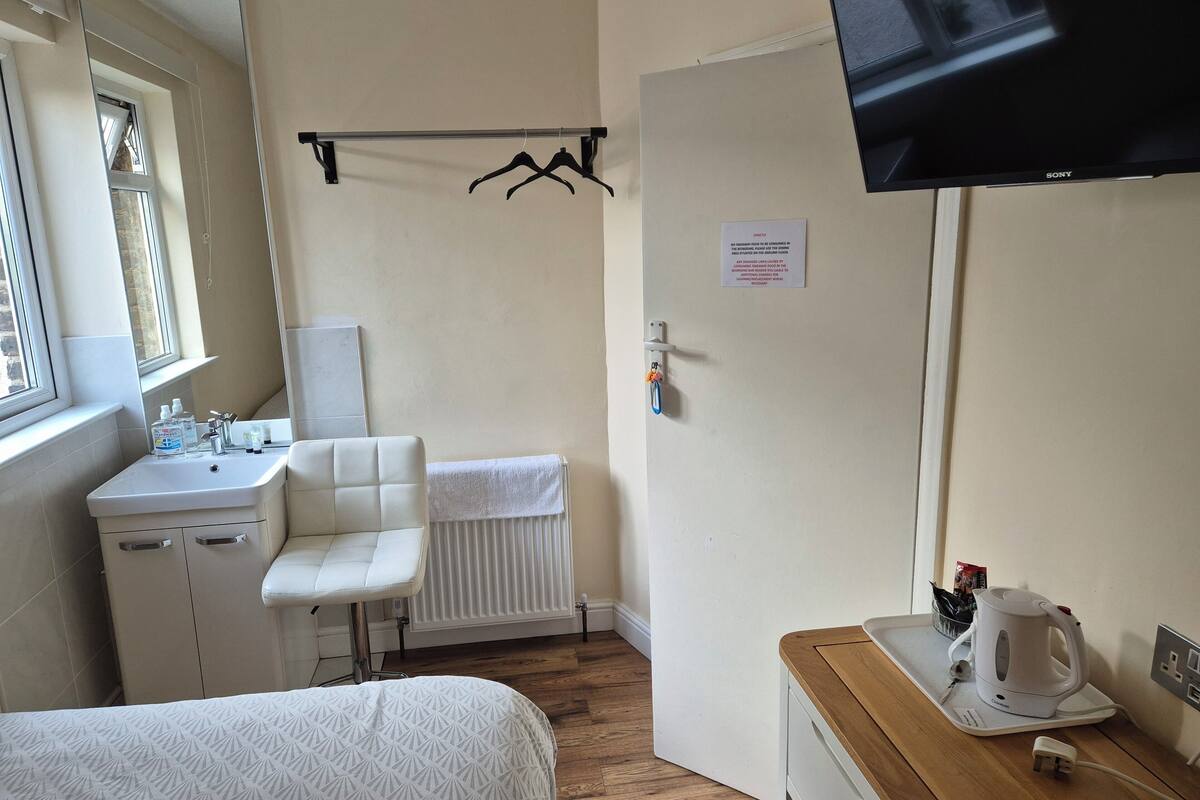 A hotel room with a bed, a small desk with a kettle, a radiator, a sink, and a TV mounted on the wall.