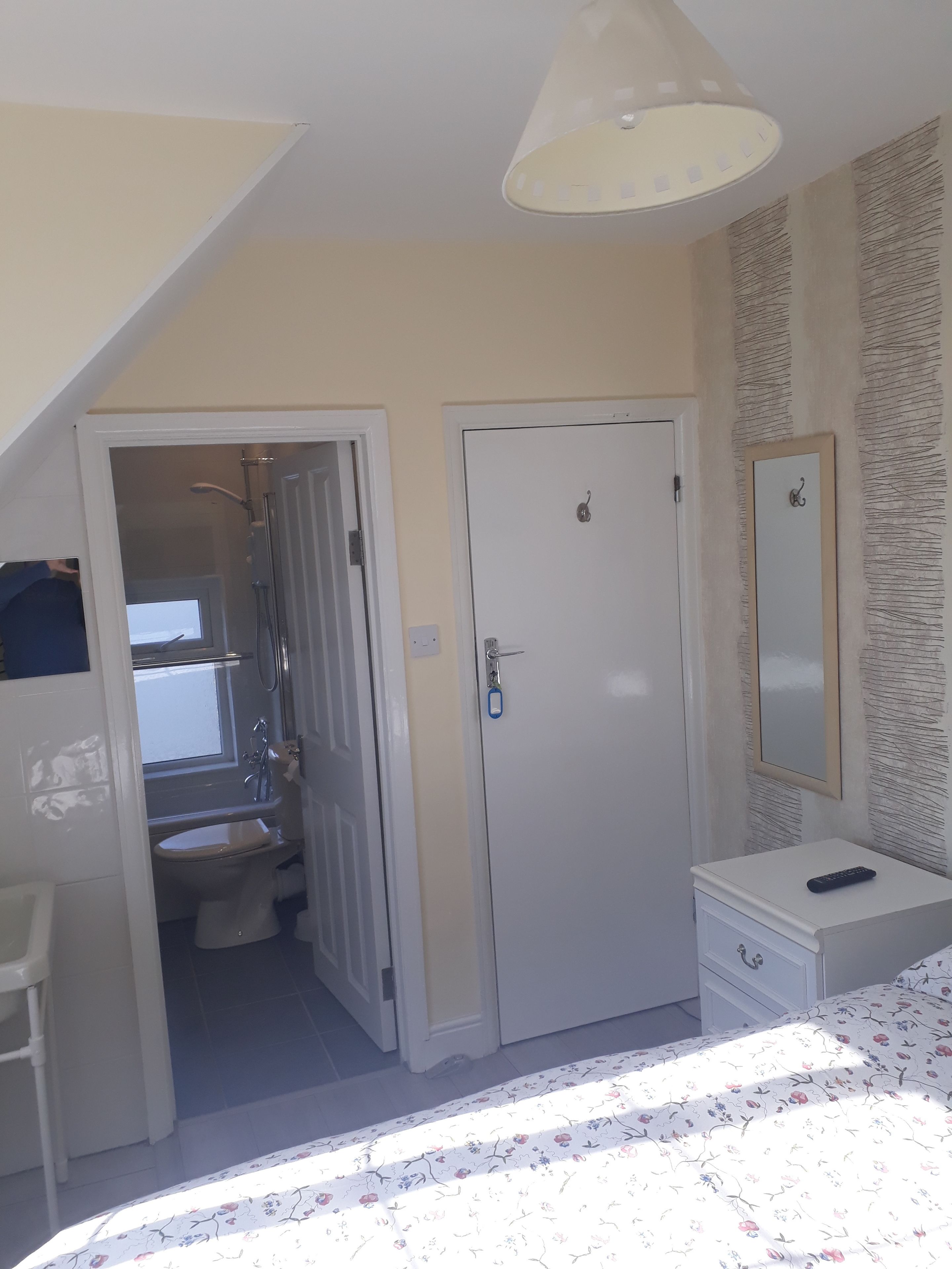 Une chambre à coucher comprenant un lit, une table de chevet, un miroir, une armoire et une salle de bain visible à travers une porte ouverte.