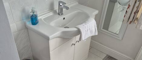 A bathroom with a white sink, mirror, and tiled walls. There is a shelf above the sink with toiletries, and a towel hangs on the side.