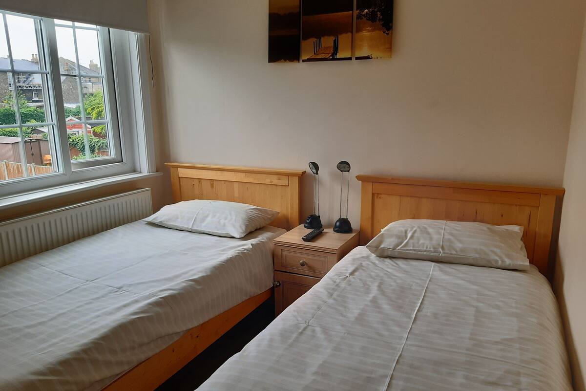 A bedroom with two single beds, wooden headboards, a nightstand with a lamp, and a view of the outside through a window.
