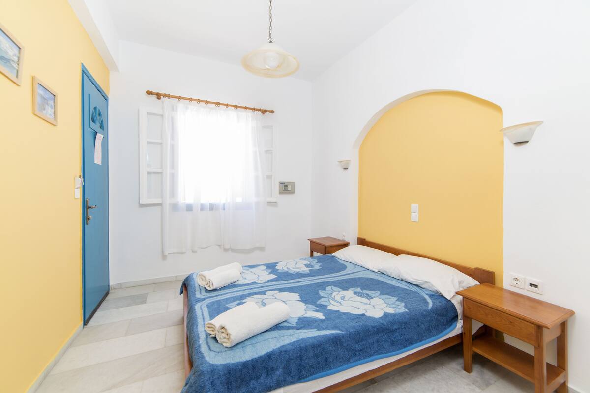 A bedroom with a bed, a nightstand, a wooden headboard, a window with curtains, and a door.