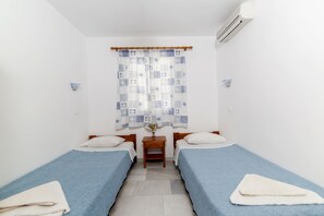 A small, clean room with two single beds, a wooden nightstand, a small table with a vase, and a window with a patterned curtain.