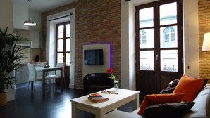 A modern living room with a white table, a black chair, and a sofa with orange cushions. There is a TV mounted on the brick wall and a window with a view of the outside. - Apartamentos Turisticos Mauror (Granada)