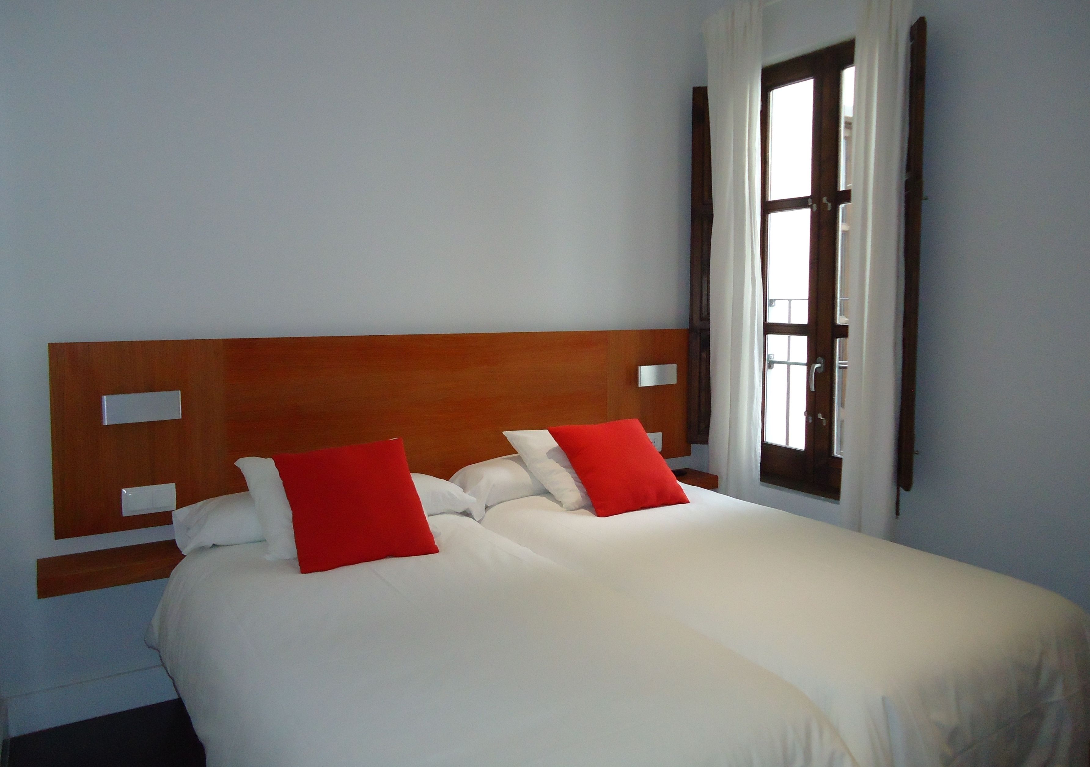 A double bed with white and red pillows, a wooden headboard, and a window with curtains.