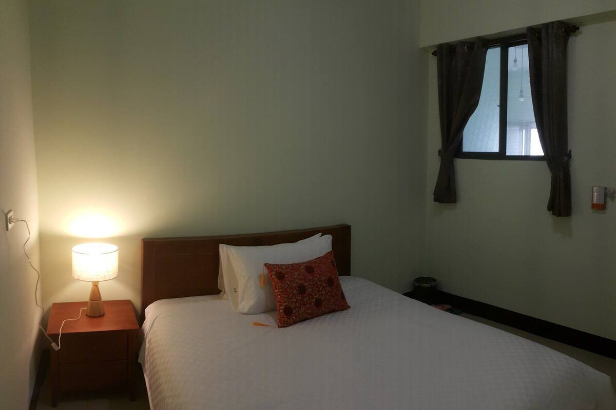 A hotel room with a bed, a nightstand, a lamp, a window with curtains, and a wall-mounted heater.