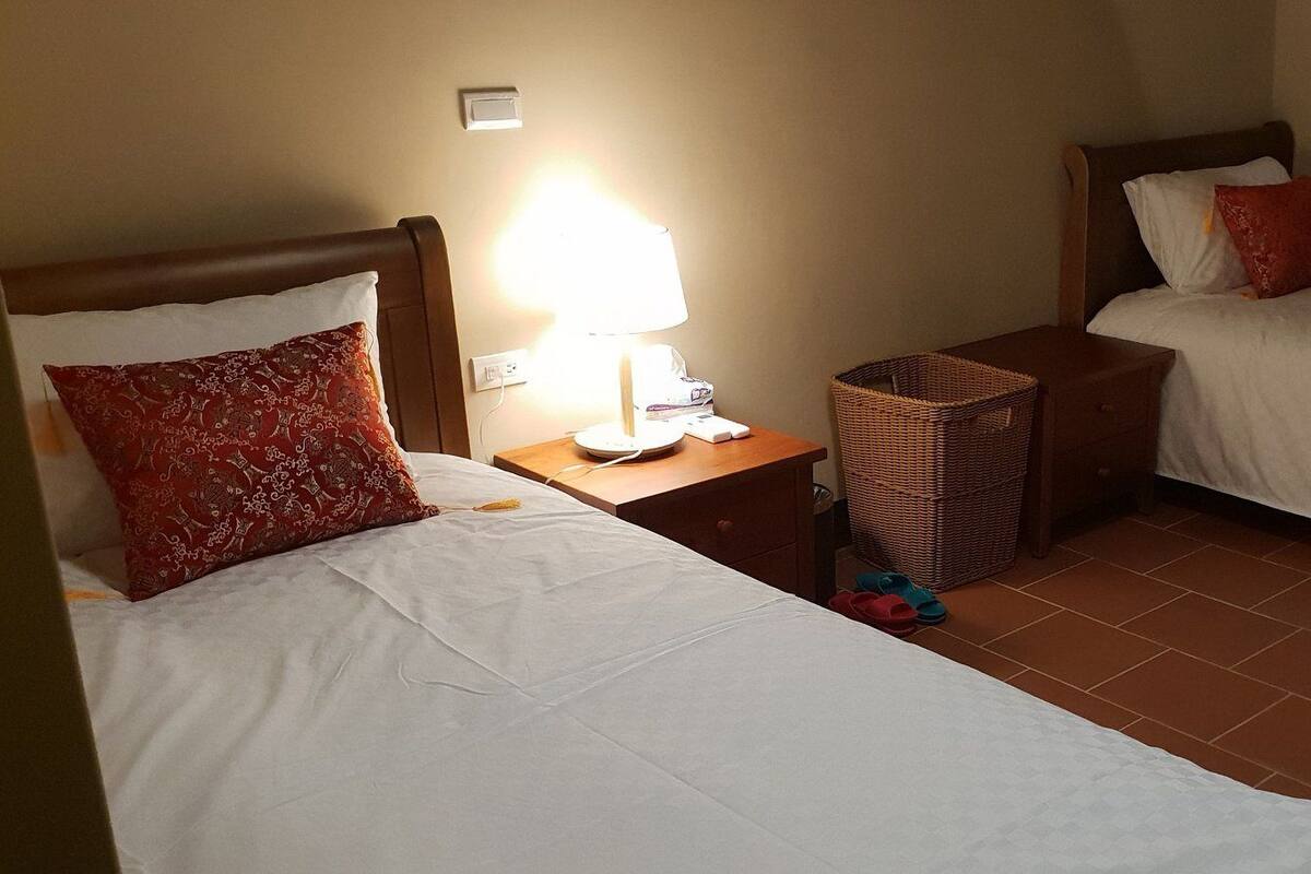 A hotel room with two beds, each with a pillow and a bedside table with a lamp. There is a wicker basket on the floor.