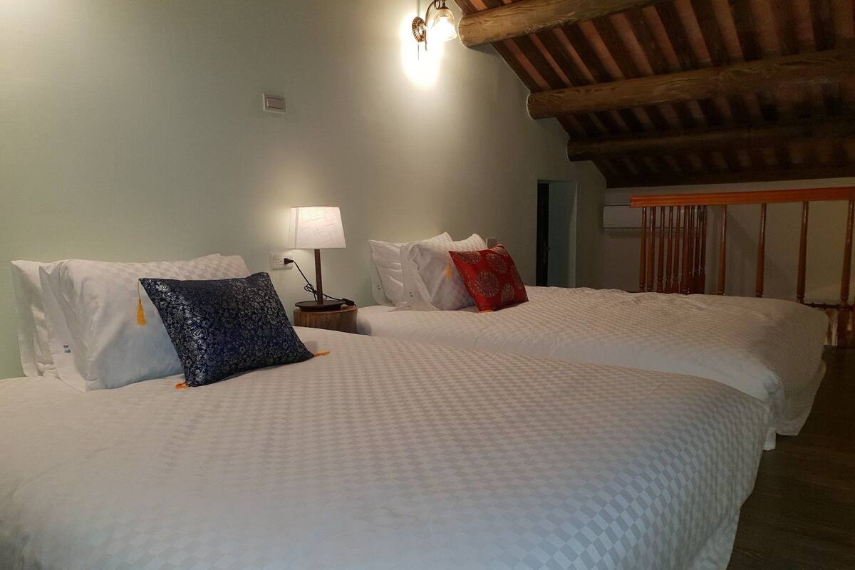 A hotel room with two beds, each with white bedding and decorative pillows. There is a wooden ceiling and a wall-mounted lamp.