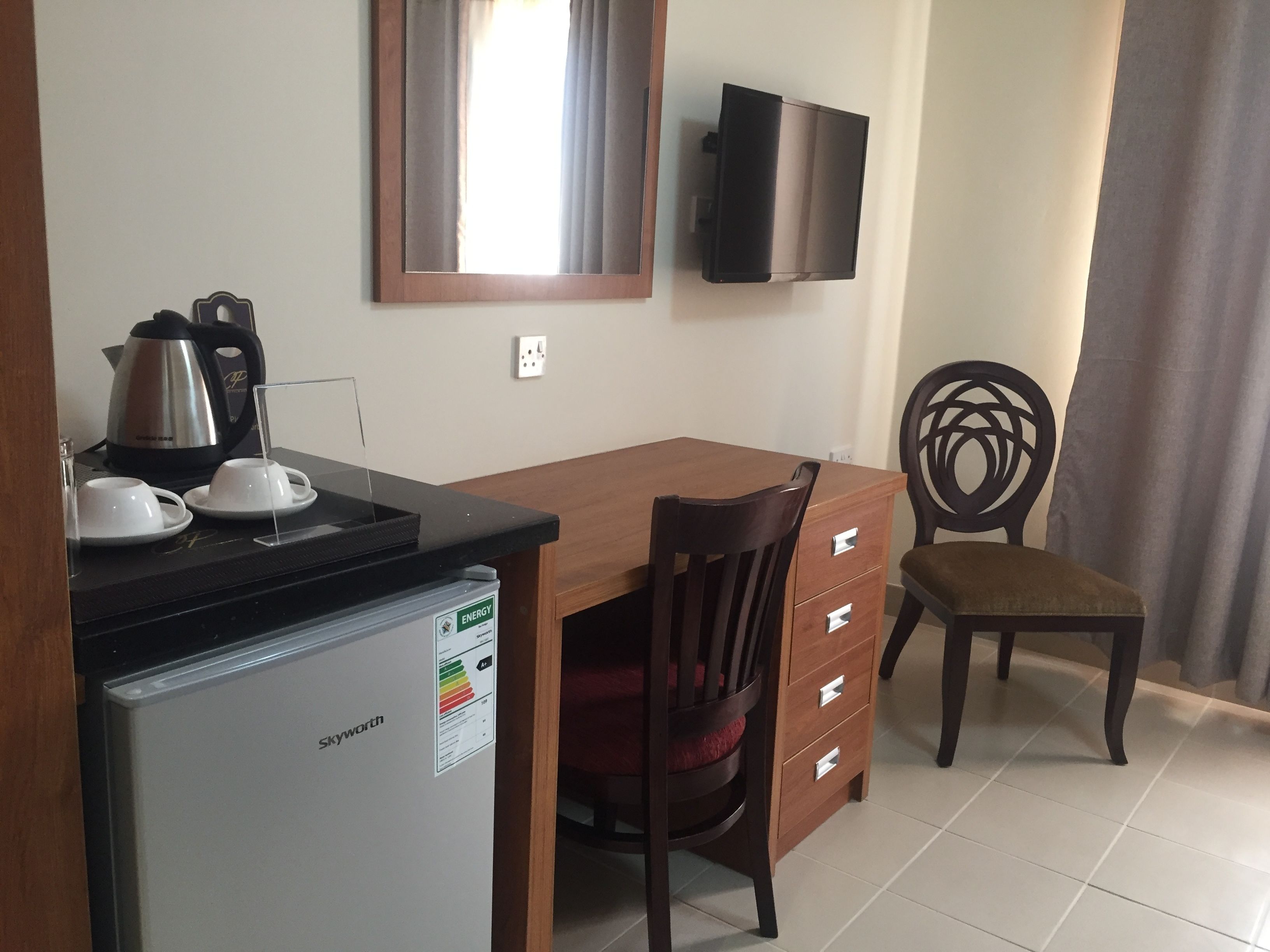 A compact kitchen with a refrigerator, a kettle, and a coffee maker on the counter. There is a wooden dining table with a chair and a TV mounted on the wall.