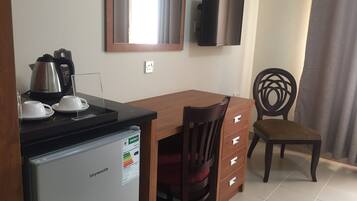 A compact kitchen with a refrigerator, a kettle, and a coffee maker on the counter. There is a wooden dining table with a chair and a TV mounted on the wall.