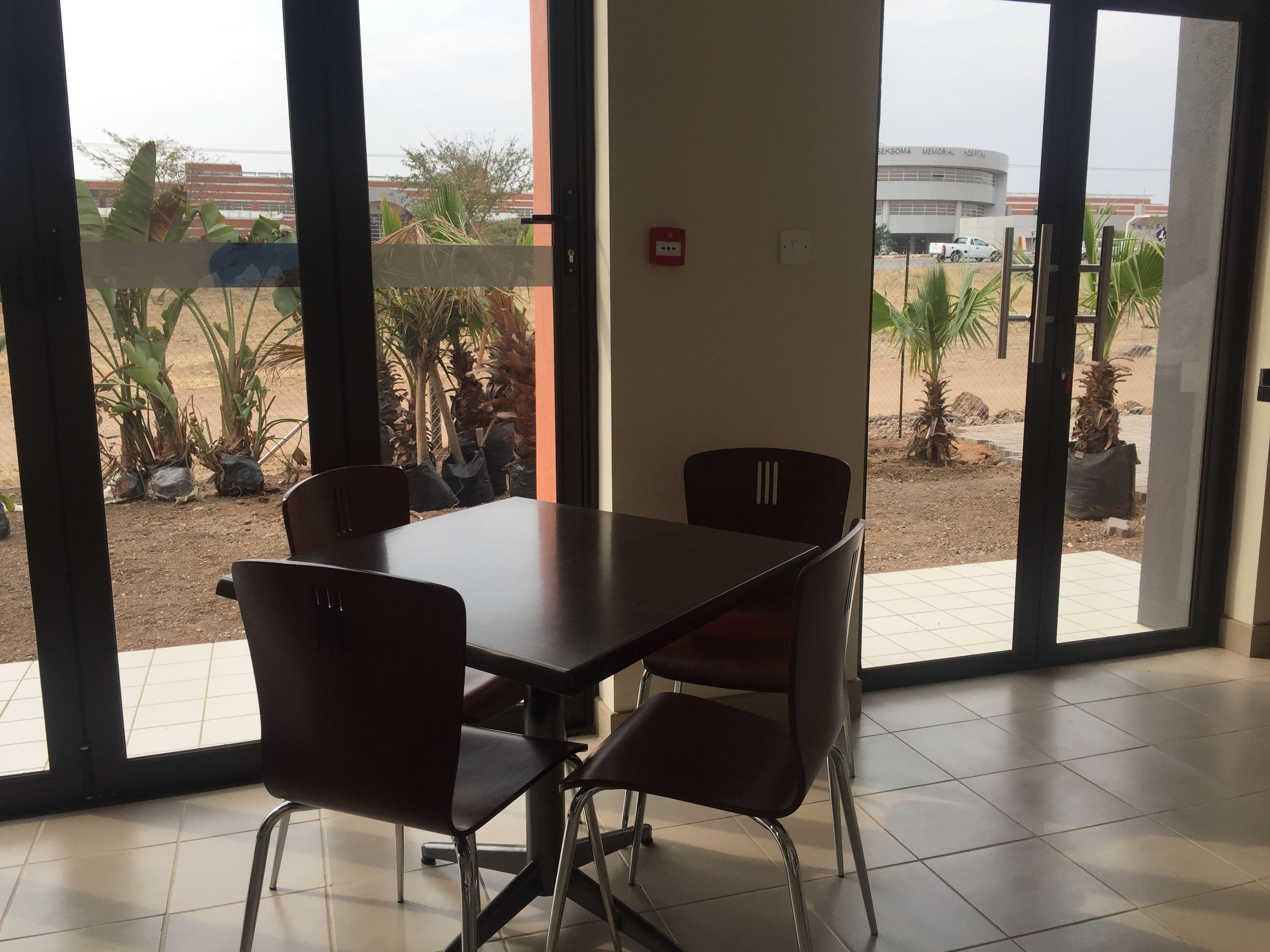 Una habitación con una mesa y sillas, ventanas amplias que dan a vistas de un edificio y palmeras.