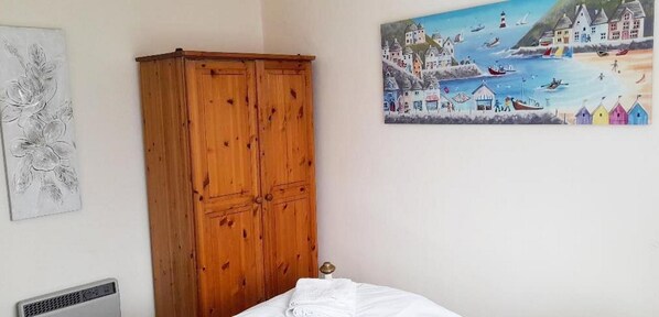 Une chambre avec une armoire en bois, un lit avec une literie blanche, un radiateur et un tableau représentant un paysage marin accroché au mur.