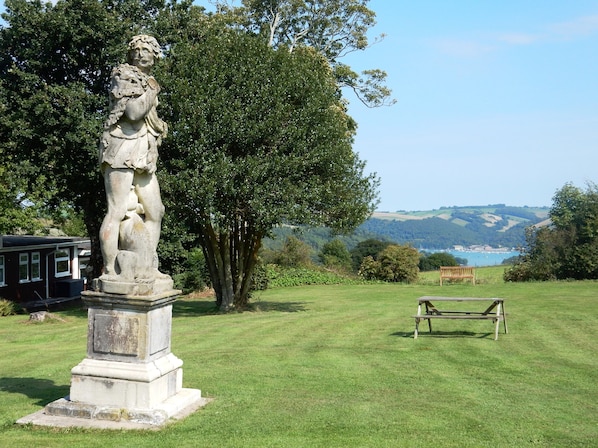 A statue in a grassy area with a bench and a view of hills and water in the background.