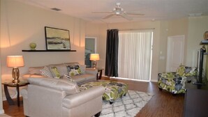 A living room with a sofa, two lamps, a ceiling fan, a large window with curtains, and a television. - Ip60503 - The Shire at West Haven - 4 Bed 3 Baths Villa (Davenport)