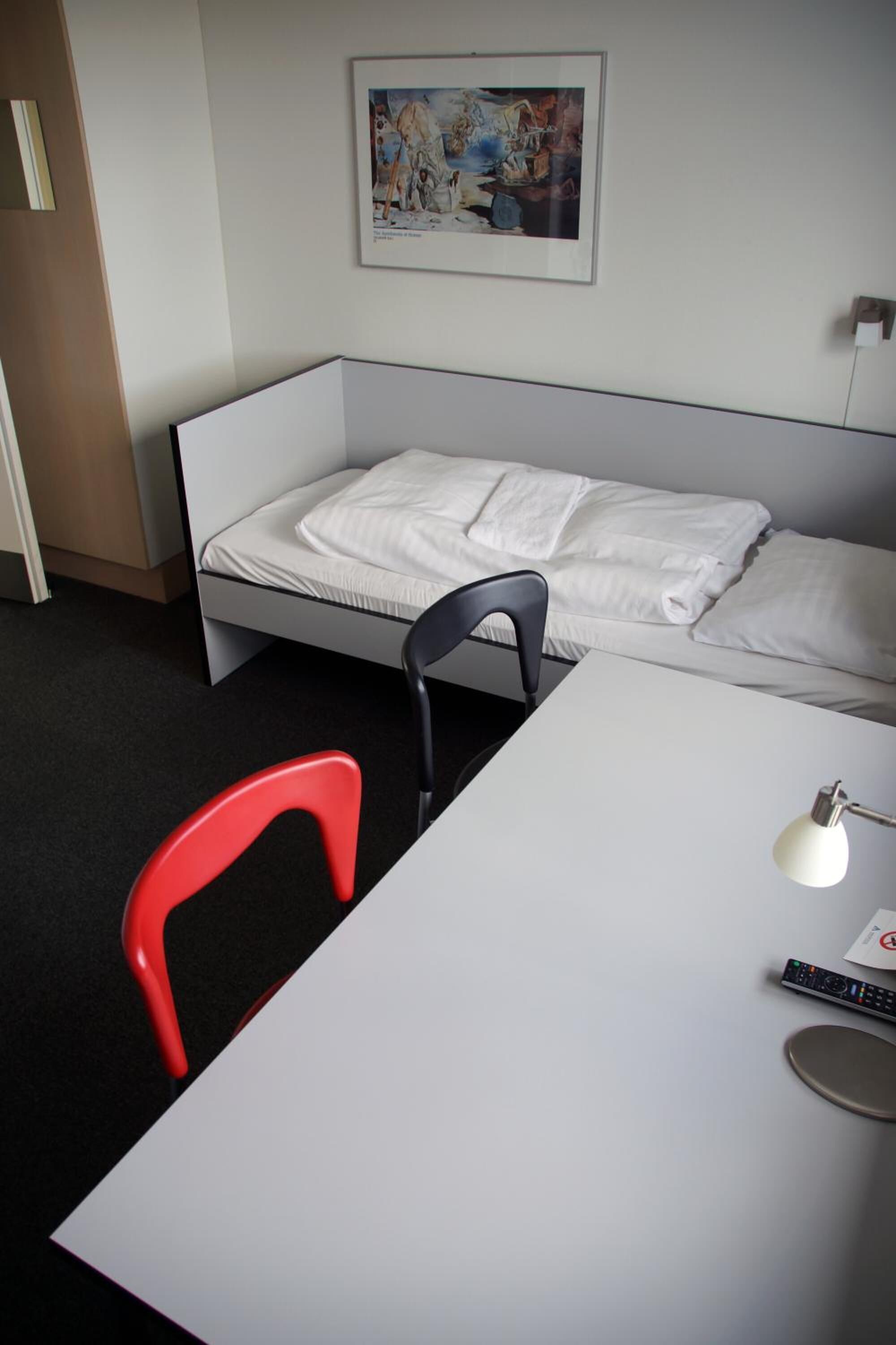 Ein Einzelbett mit weißer Bettwäsche, ein roter Sessel, ein weißer Schreibtisch, eine Lampe und eine Fernbedienung.