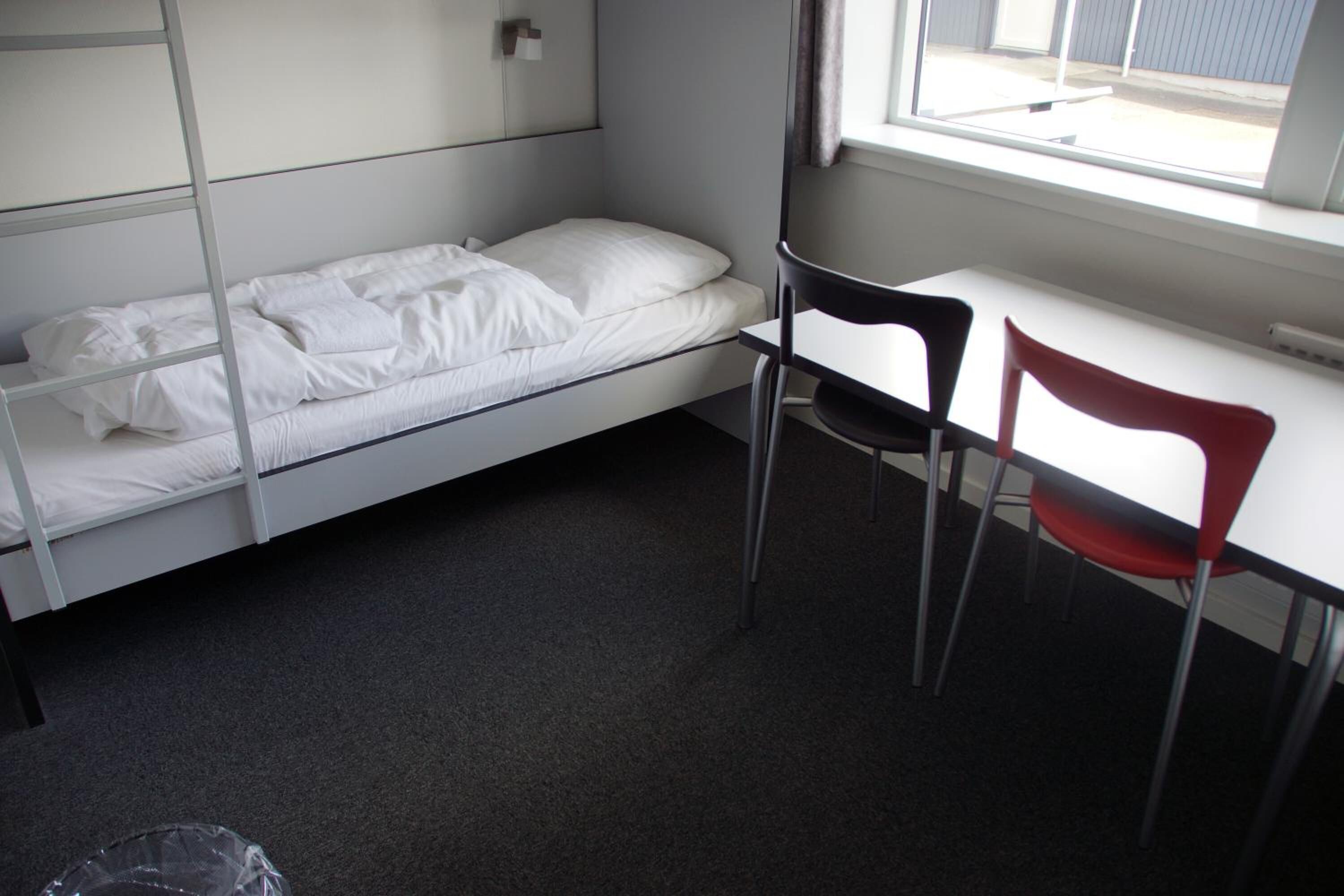 Una habitación pequeña con cama individual, escritorio y dos sillas, una roja y otra negra.