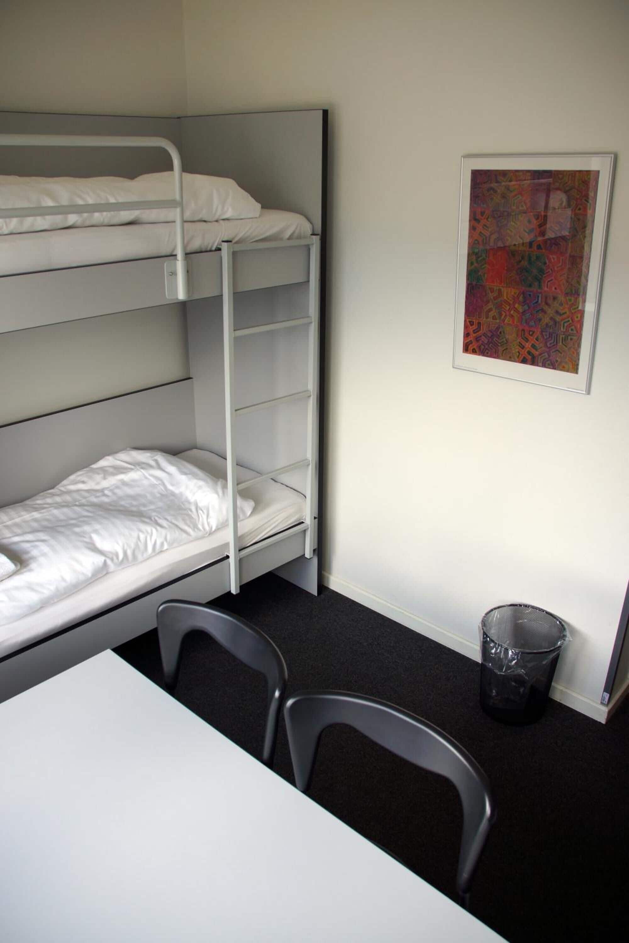 Une chambre avec un lit superposé, une table et des chaises, une œuvre d’art encadrée au mur et une poubelle.