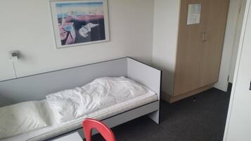 A single-bed room with a white mattress, a red chair, a wooden wardrobe, and a painting on the wall.