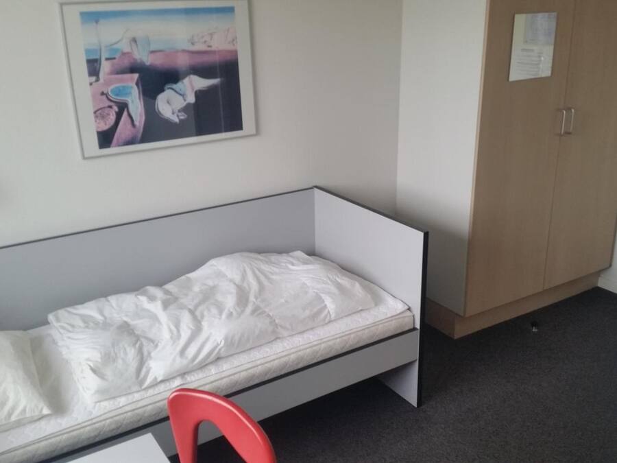 A single-bed room with a white mattress, a red chair, a wooden wardrobe, and a painting on the wall.