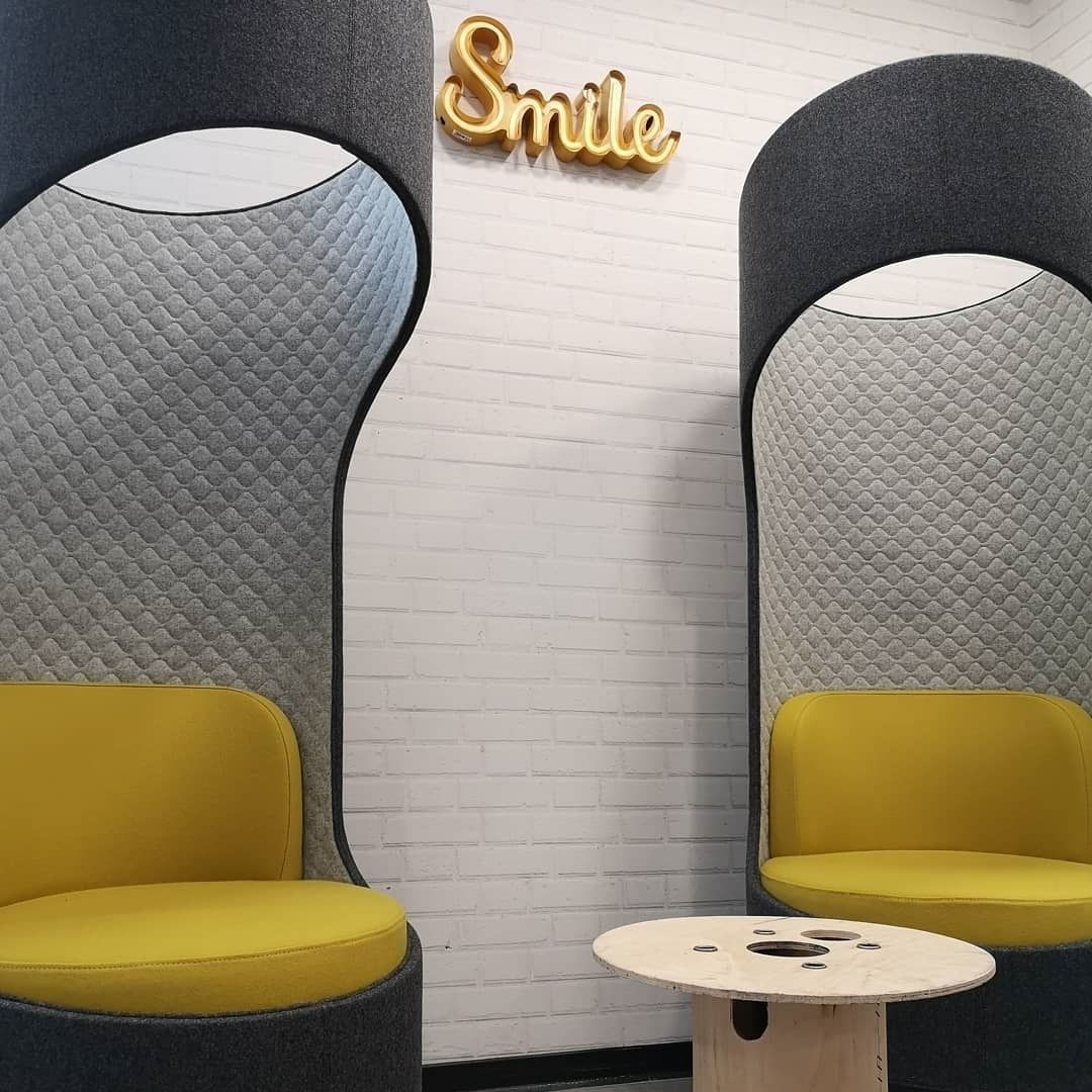 Twee moderne stoelen met gewatteerde rugleuningen en gele kussens, een klein houten tafeltje, en een muur met een 'Smile'-bord.