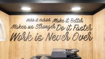 A room with two large black printers, a wooden wall with motivational text, and a ceiling with a grid pattern and recessed lighting.