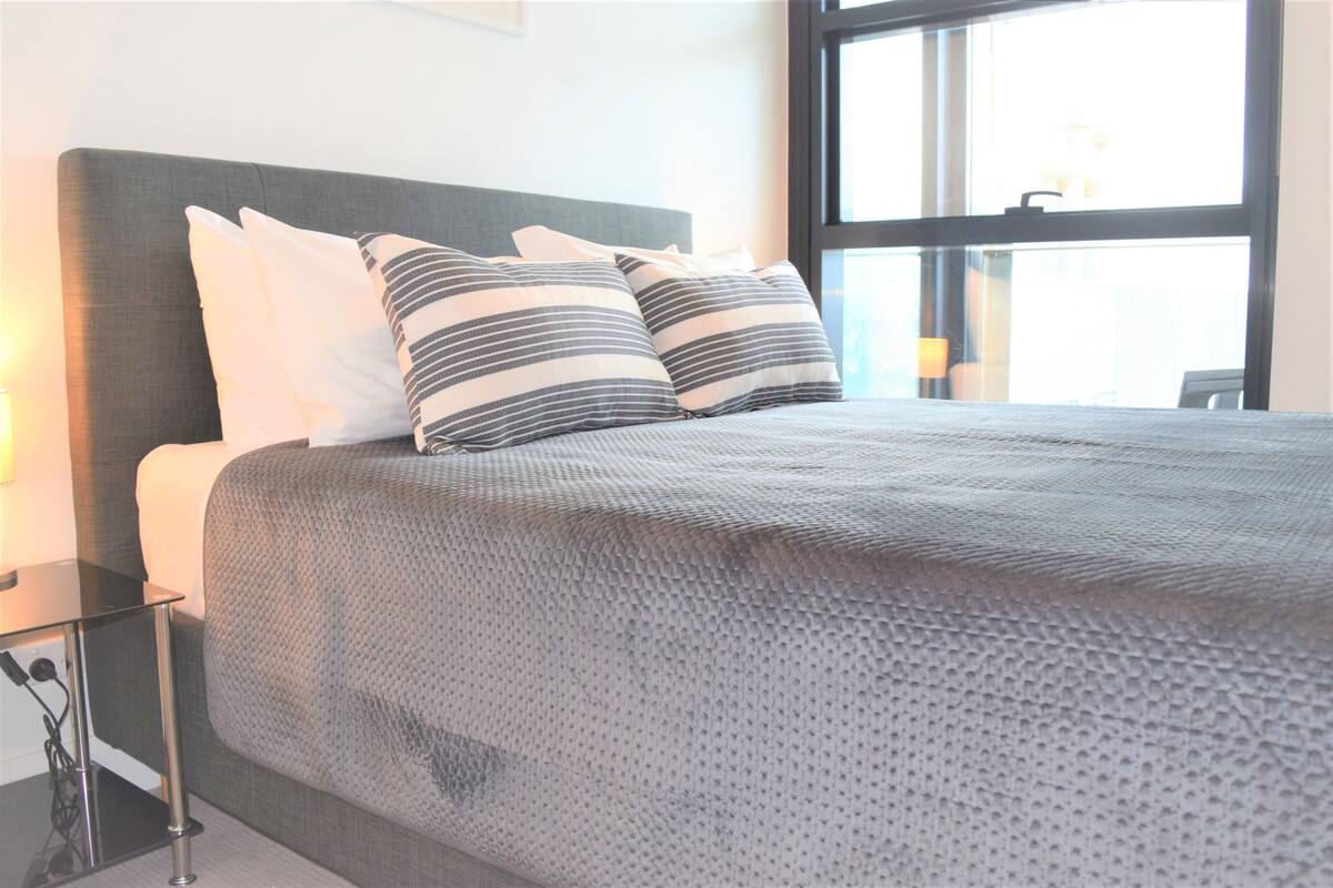 Una cama bien hecha con una colcha gris texturizada y almohadas a rayas, una mesita de noche con una lámpara, y un ventanal grande que proporciona luz natural.