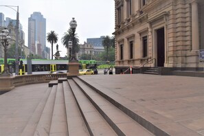 Una plaza pública con escalones que conducen a la entrada de un edificio, una estatua y un autobús verde al fondo.
