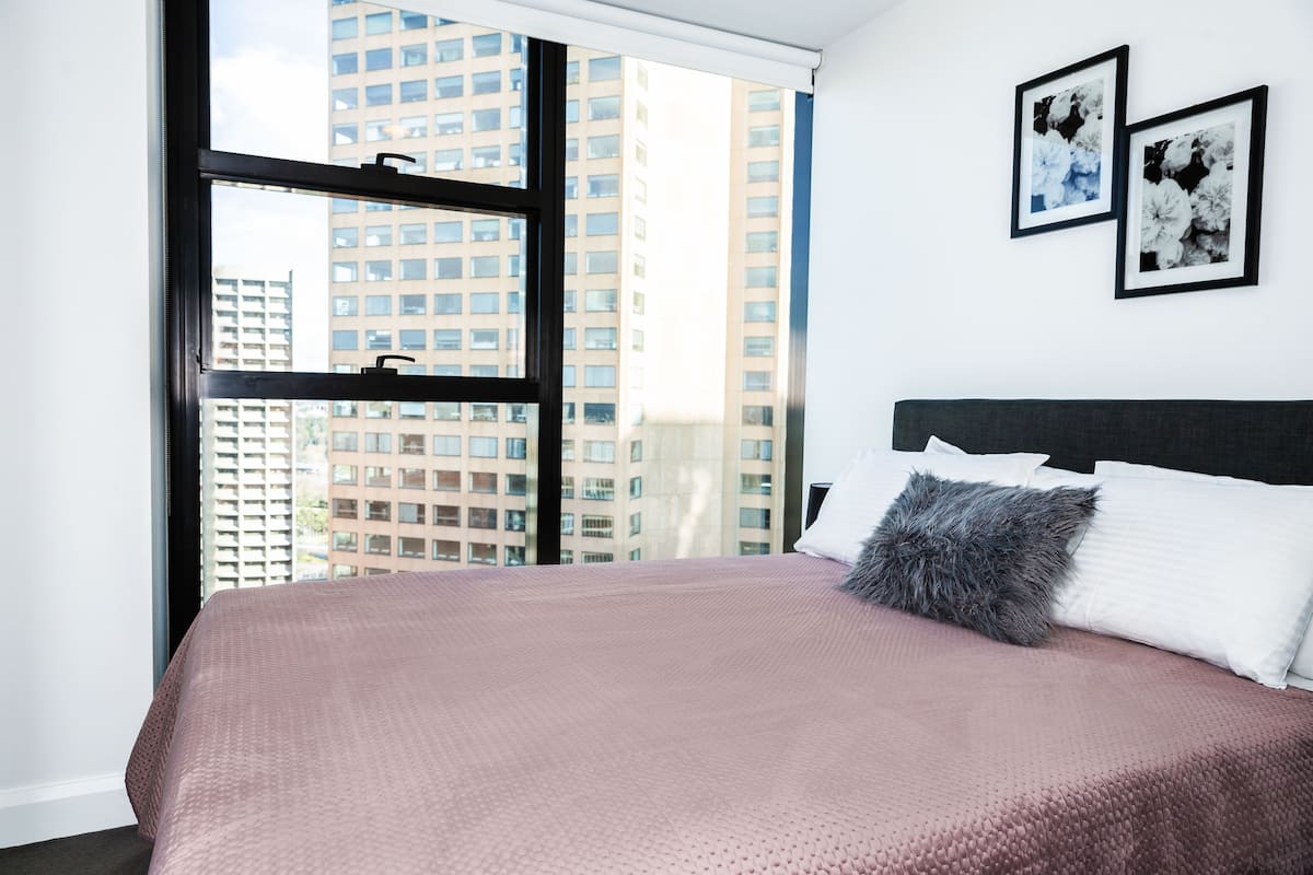 Un dormitorio con un ventanal grande, una cama con colcha rosa, almohadas blancas y dos cuadros en la pared.