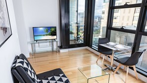A modern living room with a sofa, a glass table with chairs, a TV on a stand, and large windows offering a city view. - Executive Stay on Little Collins (Melbourne)