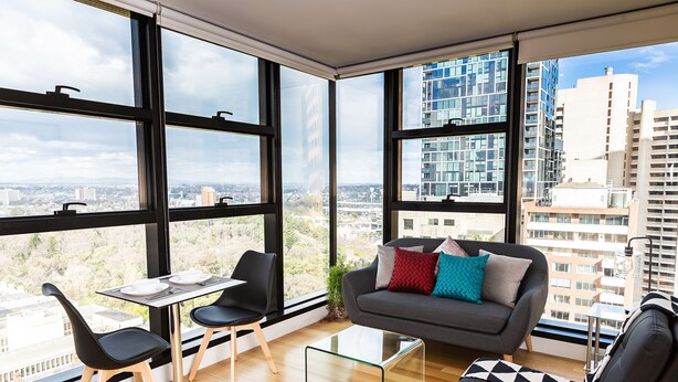 A modern living room with large windows, a sofa, two chairs, a coffee table, and a small table with a laptop. - Executive Stay on Little Collins (Melbourne)