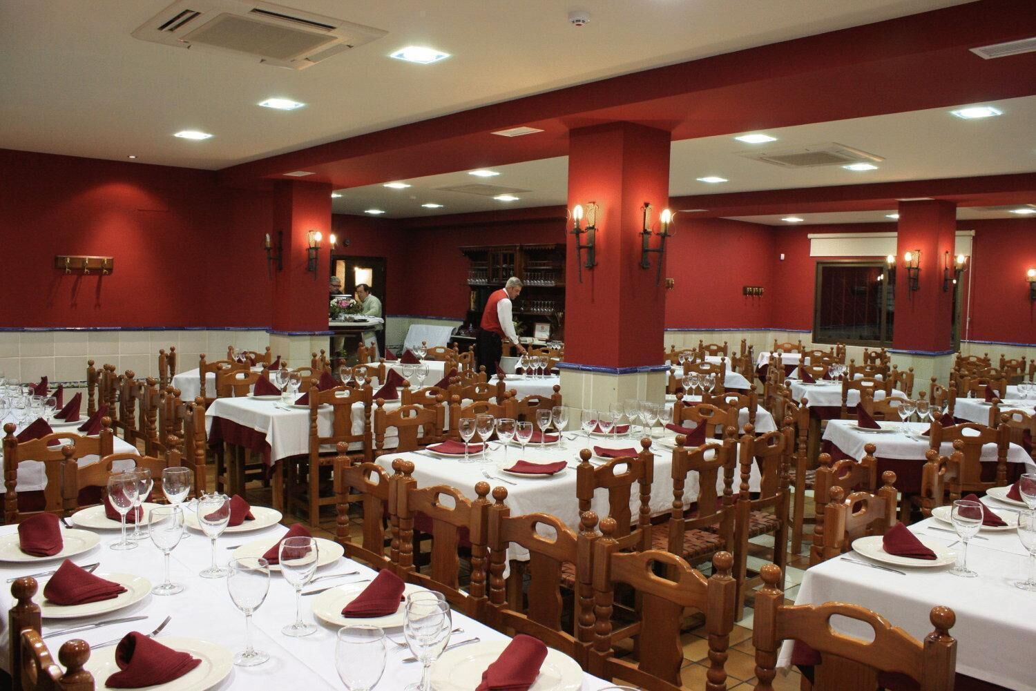 Une grande salle à manger avec de nombreuses tables dressées pour le service, des chaises en bois et des serviettes rouges.
