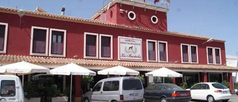 Un edifici vermell amb un rètol que diu "Hostal Restaurante La Bartola" i diversos cotxes aparcats al davant.