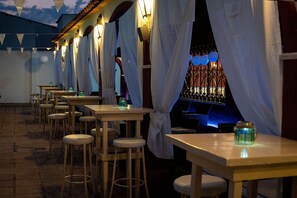 An outdoor restaurant with wooden tables and chairs, white curtains, and a view of a building with a blue-lit entrance. - Hostal La Bartola (Córdoba)