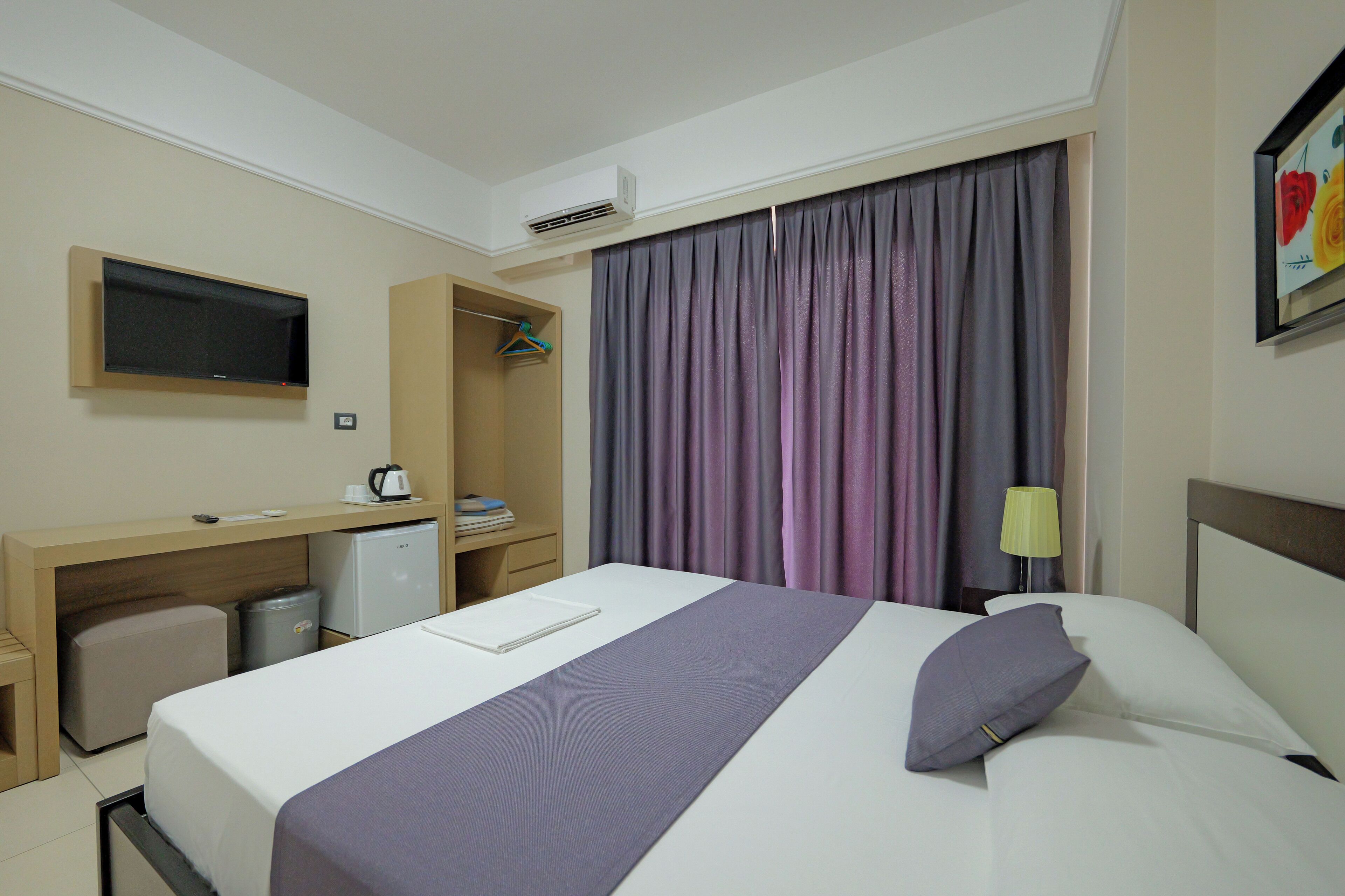 A hotel room with a bed, a desk, a TV, a mini-fridge, a lamp, and a window with curtains.