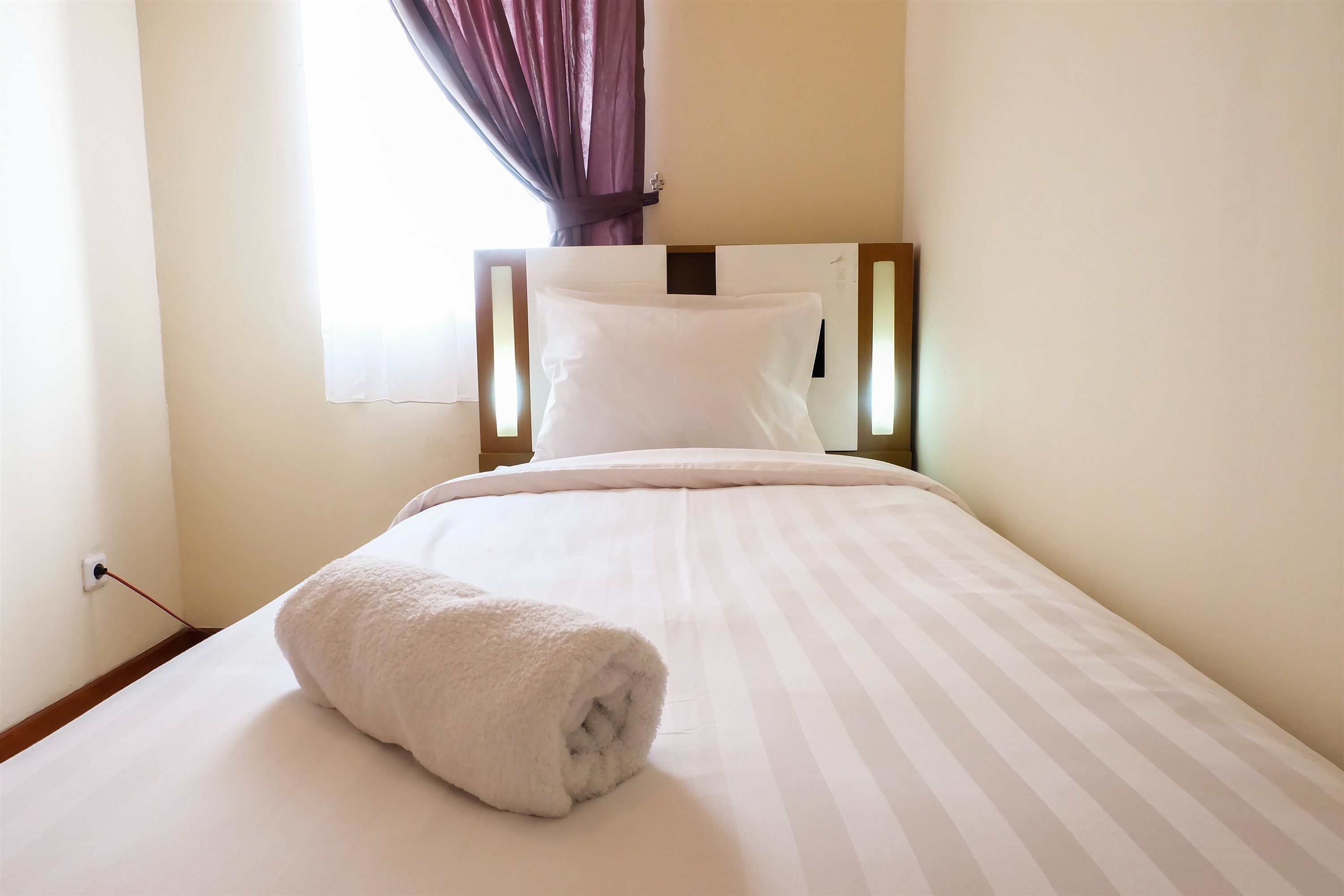 A neatly made bed with a rolled white towel, a wooden headboard, and a window with curtains.