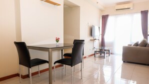 A modern living room with a glass dining table, black chairs, a grey sofa, a TV, and a window with curtains. - Grand Palace Kemayoran Apartment For Lifestyle Living (Jakarta)