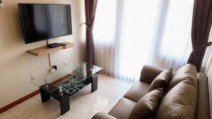 A modern living room with a flat-screen TV mounted on the wall, a glass coffee table, a beige sofa, and sheer curtains allowing natural light. - Grand Palace Kemayoran Apartment For Lifestyle Living (Jakarta)
