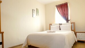 A neatly made bed with white linens and two pillows, a wooden nightstand with a drawer, and a window with red curtains. - Grand Palace Kemayoran Apartment For Lifestyle Living (Jakarta)