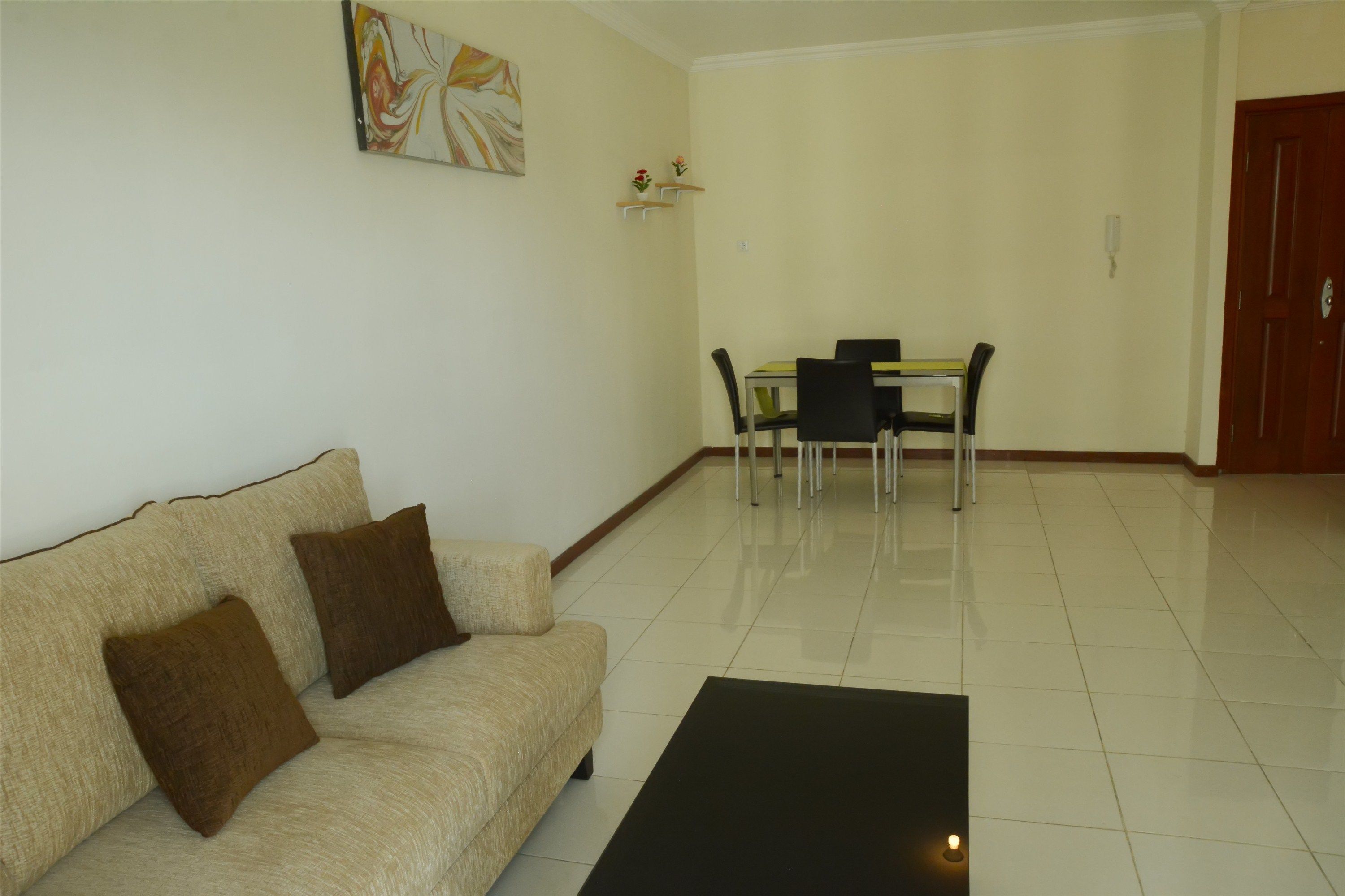 A living room with a beige sofa, two brown cushions, a black coffee table, a dining table with four chairs, and a wooden door.