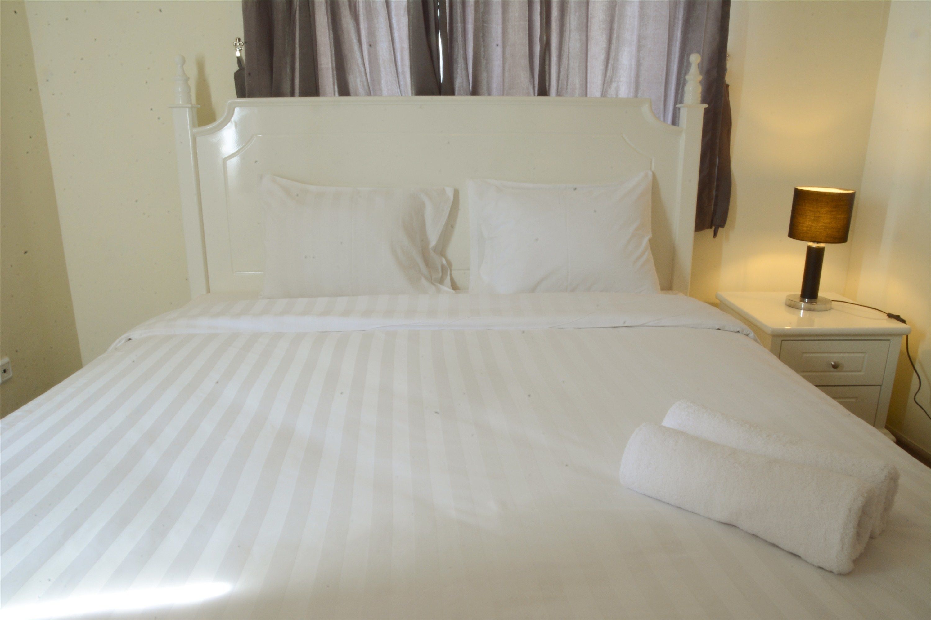 A neatly made bed with white bedding and pillows, a bedside lamp, and a small bedside table with a drawer.