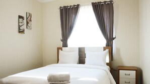 A neatly made bed with white linens and a folded towel, a wooden nightstand with drawers, and two abstract paintings on the wall. - New And Modern Grand Palace Kemayoran Apartment (Jakarta)