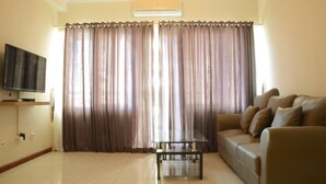 A living room with a flat-screen TV, a glass coffee table, a sofa, and a large window with curtains. - New And Modern Grand Palace Kemayoran Apartment (Jakarta)