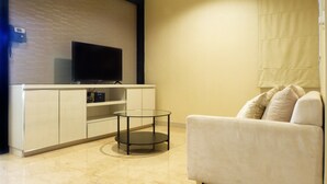 A modern living room with a flat-screen TV mounted on a white cabinet, a glass coffee table, and a white sofa with cushions. - Best North L'Avenue Apartment (Jakarta)
