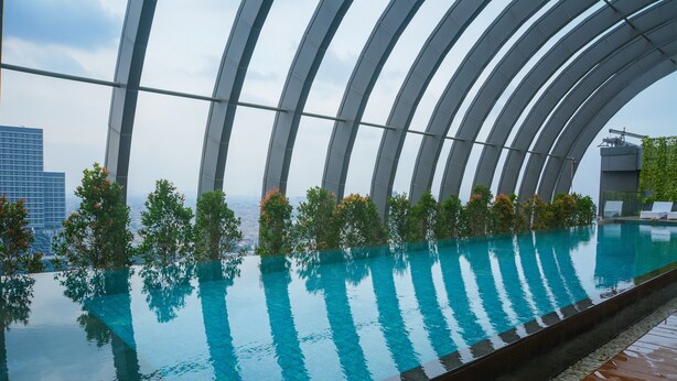 An infinity pool with a clear blue water surface, surrounded by a modern architectural structure with curved metal beams. - Best North L'Avenue Apartment (Jakarta)