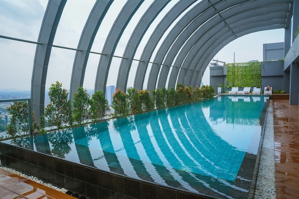 Una piscina infinita en la azotea con techo de vidrio curvo, rodeada de vegetación y vistas a la ciudad.