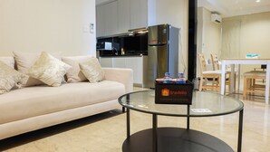 A modern living room with a glass coffee table, a white sofa with patterned cushions, a kitchen area with a stainless steel refrigerator, and a dining table with chairs. - Best North L'Avenue Apartment (Jakarta)