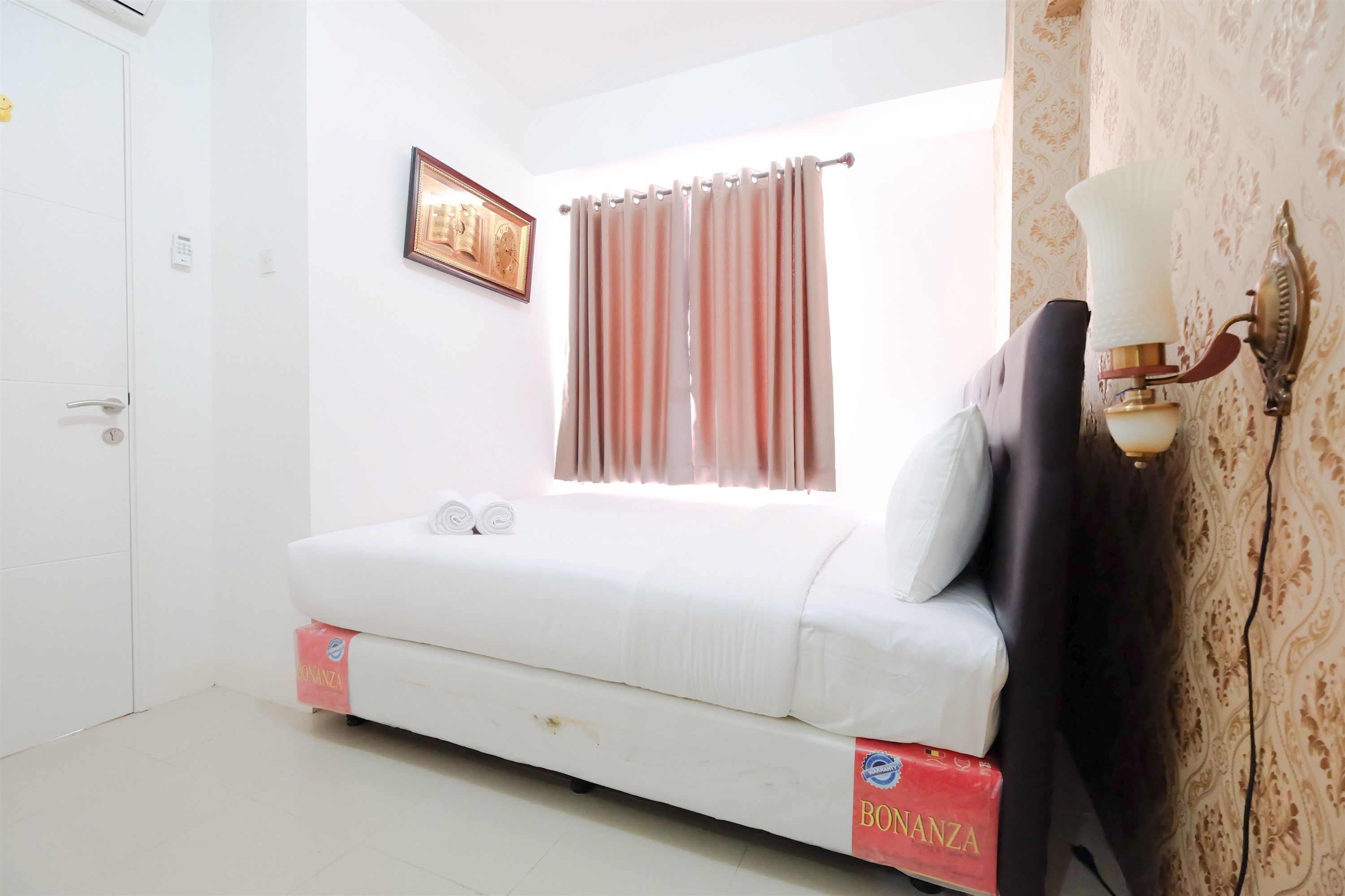 A bedroom with a bed, a window with curtains, a wall-mounted lamp, and a door.