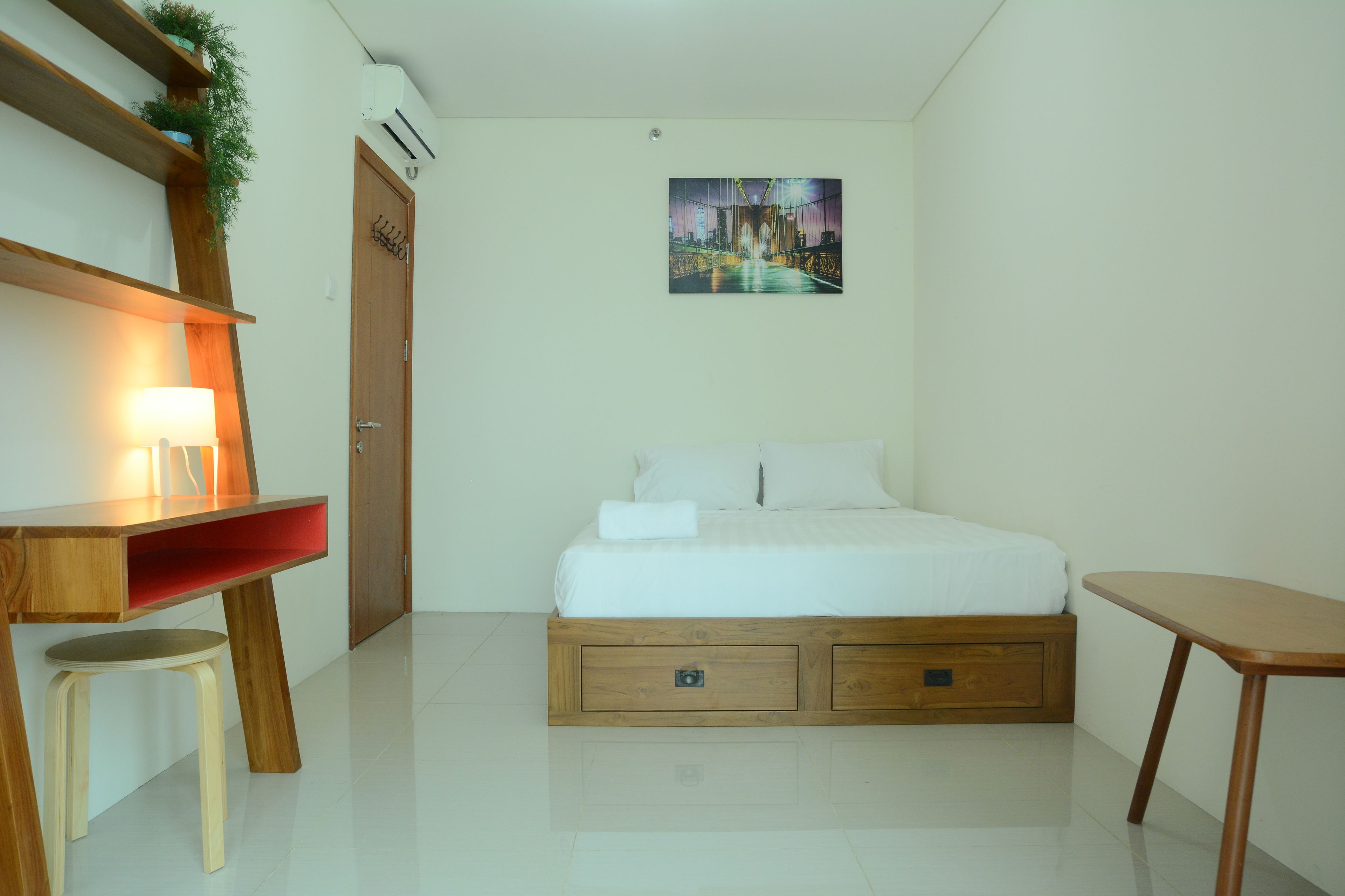 A bedroom with a bed, a desk with a chair, a small stool, a wooden nightstand, and a wall-mounted air conditioner.