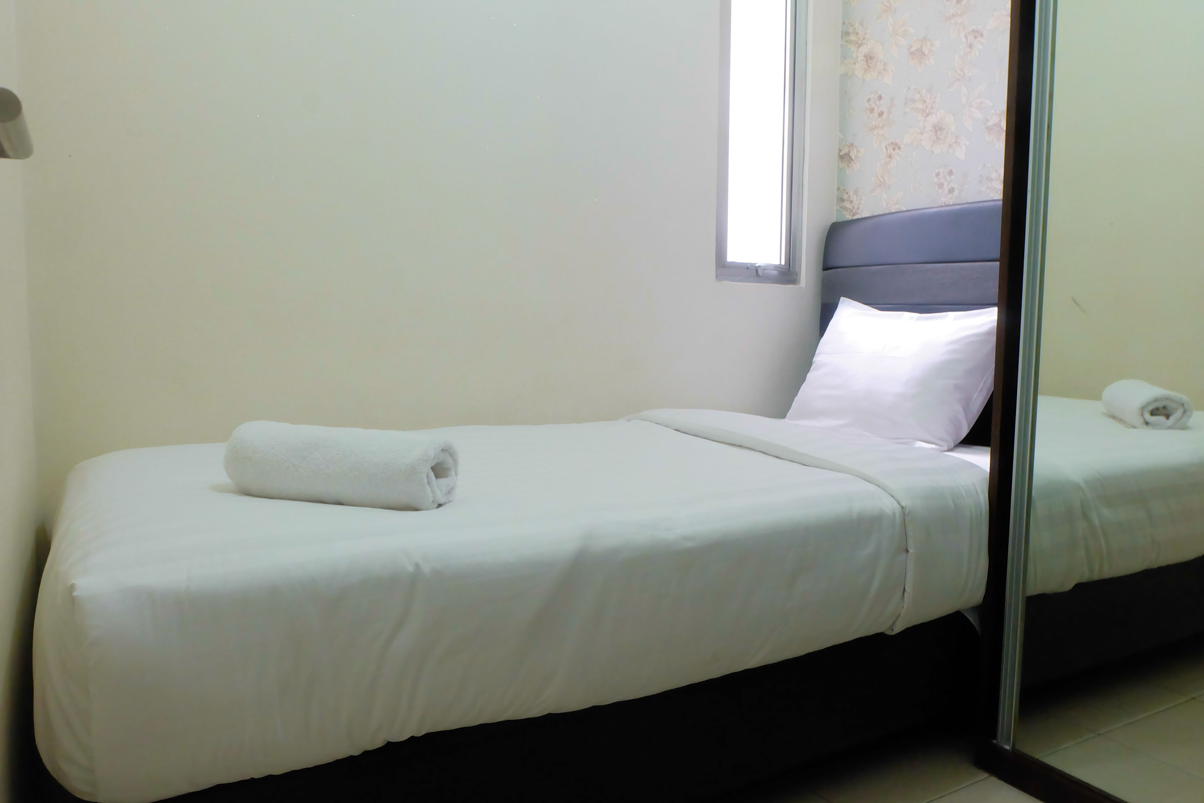A single bed with white bedding and a rolled towel, a window with floral wallpaper, and a mirror reflecting part of the room.