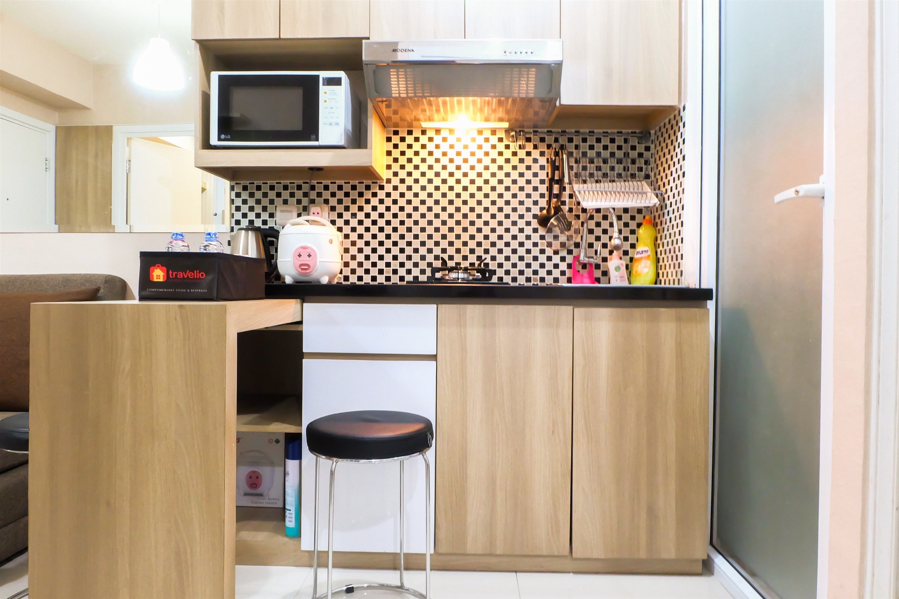 A compact kitchen with a microwave, a gas stove, and a sink. There is a bar stool and a small refrigerator.