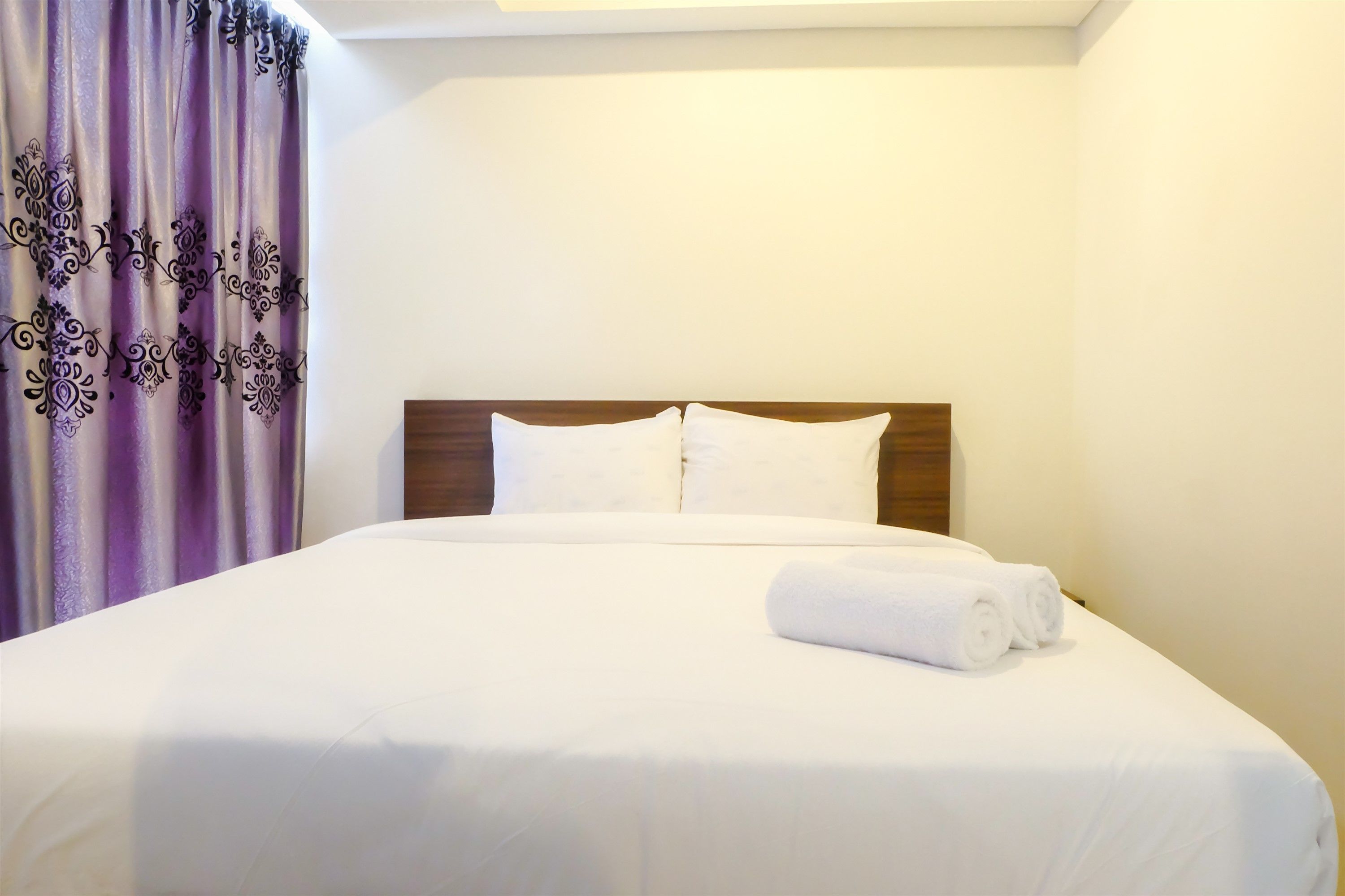 A neatly made bed with white linens and pillows, a wooden headboard, and a purple curtain with a paisley pattern.